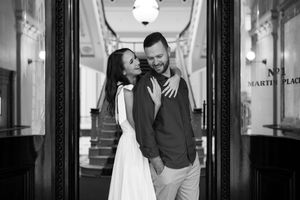 Black and white photo of a smiling couple embracing in an elegant hallway doorway, woman in white dress, man in casual shirt.