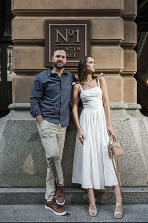 A stylish couple poses outside No.1 Martin Place, man in blue shirt and chinos, woman in white midi dress holding a handbag.