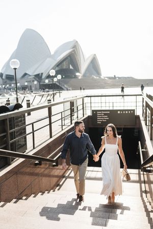 Couple holding hands walking up stairs with Sydney Opera House in the background on a sunny day.