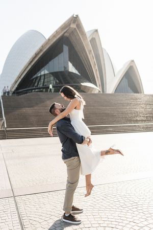 Couple embracing in front of Sydney Opera House, man lifting woman in white dress on sunny day.