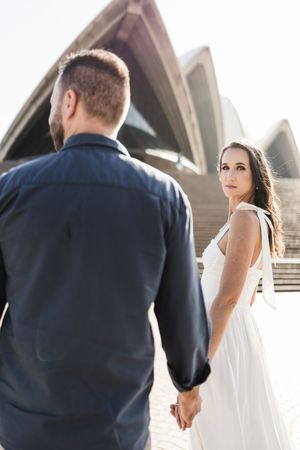 Couple holding hands in front of Sydney Opera House, bride in white dress glancing back over her shoulder.