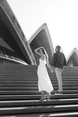 Couple holding hands on Sydney Opera House steps, woman in white dress looking back, black and white photo.