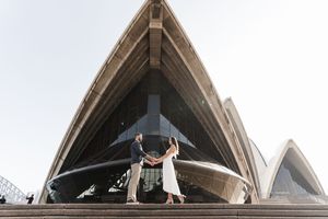 Couple holds hands on steps of Sydney Opera House, iconic shell-shaped roof soaring above them in soft natural light.