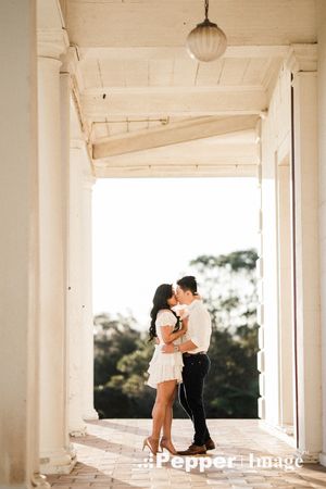 Couple sharing a romantic kiss on a sunlit colonial veranda with white columns and hanging globe light.