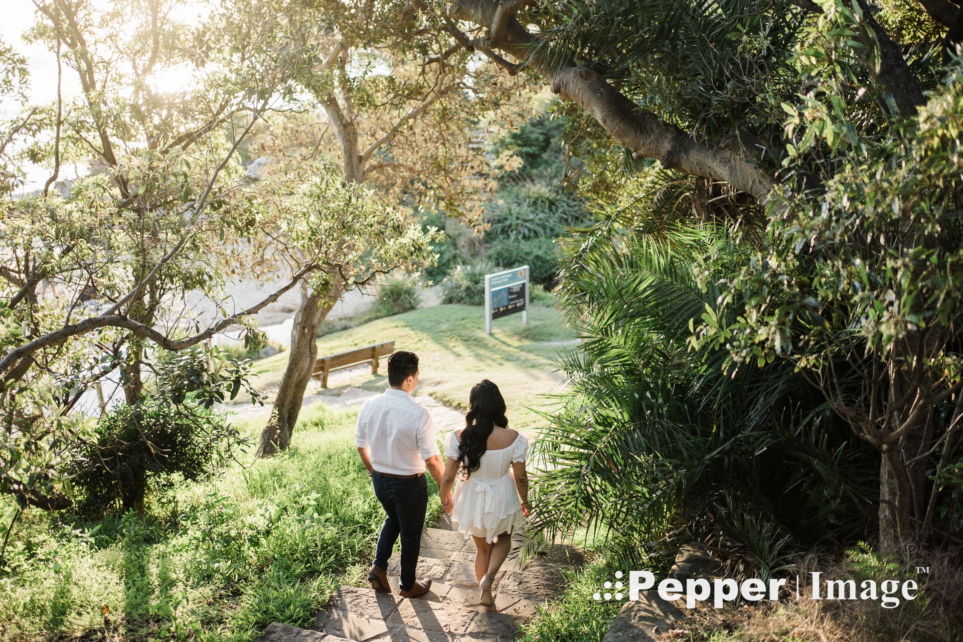 Couple walking hand-in-hand along a sunlit garden path surrounded by lush green trees and shrubs.