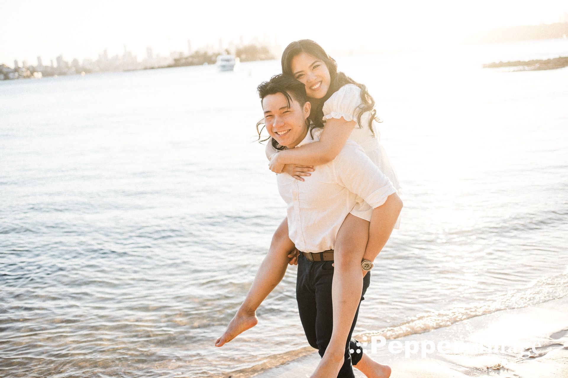 Couple laughing on beach at sunset, woman piggyback riding on mans back near sparkling ocean water.