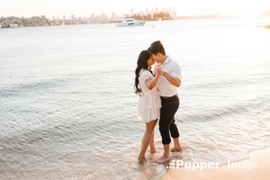 Couple sharing a romantic kiss while standing in shallow ocean water at golden hour with city skyline in background.