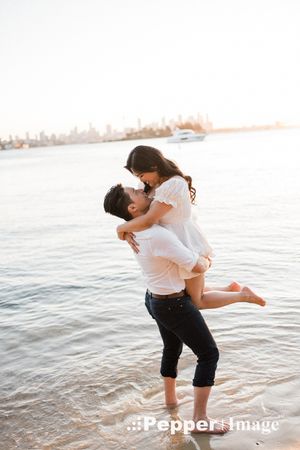 Couple sharing a romantic embrace on a sandy beach at sunset with a city skyline in the background.