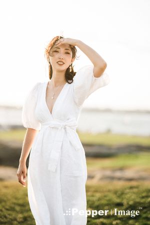 A young woman in a white wrap dress shields her eyes from the sun while standing outdoors near the coast.