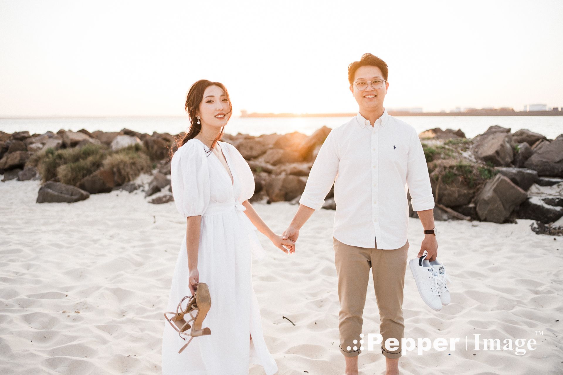 Couple holding hands on a sandy beach at sunset, wearing white outfits, with rocky shoreline in the background.