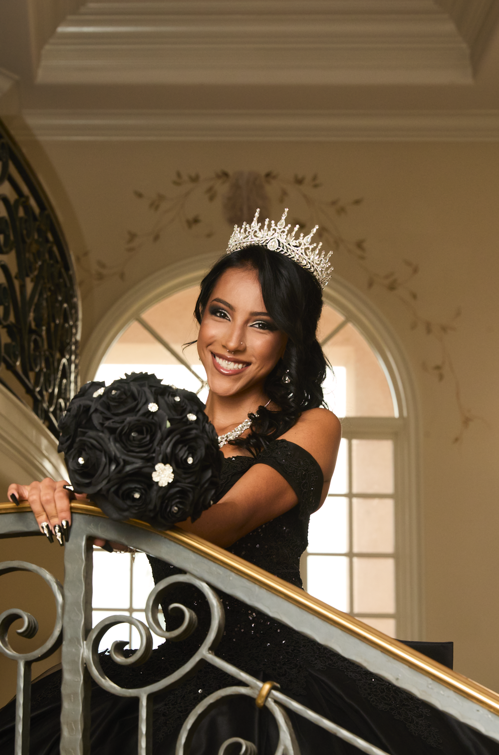 Young woman in black gown and tiara smiles holding black rose bouquet on elegant staircase.