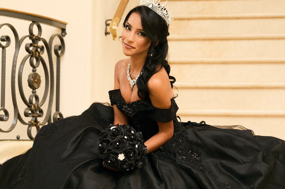 Young woman in black ball gown with tiara and bouquet poses elegantly on staircase for quinceañera.