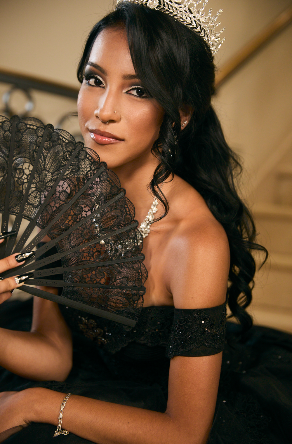 Young woman in black off-shoulder gown holding lace fan, wearing crystal tiara and jewelry, posing elegantly on staircase.
