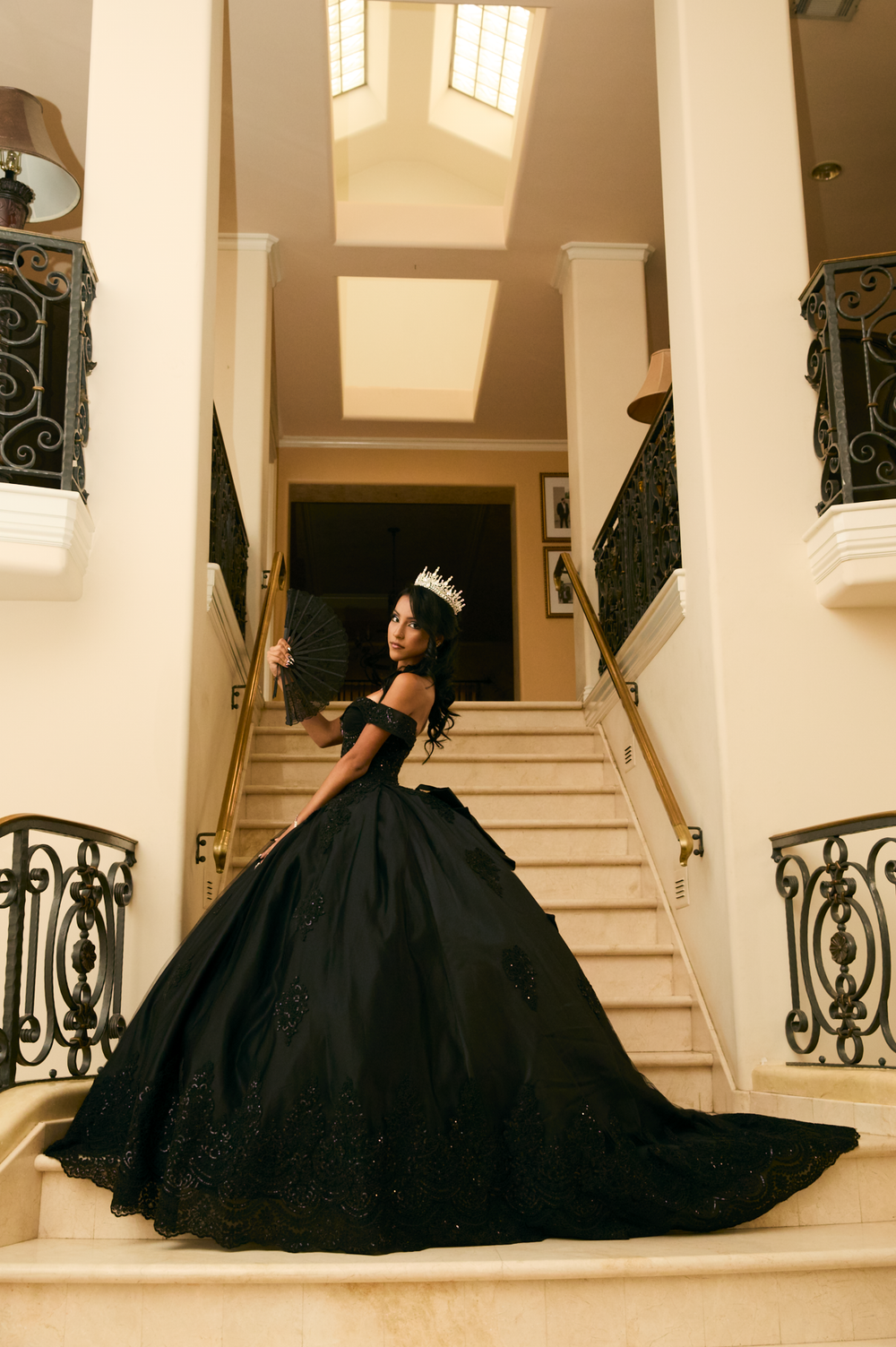 Young woman in black ball gown and crown poses elegantly on grand staircase for quinceañera celebration.