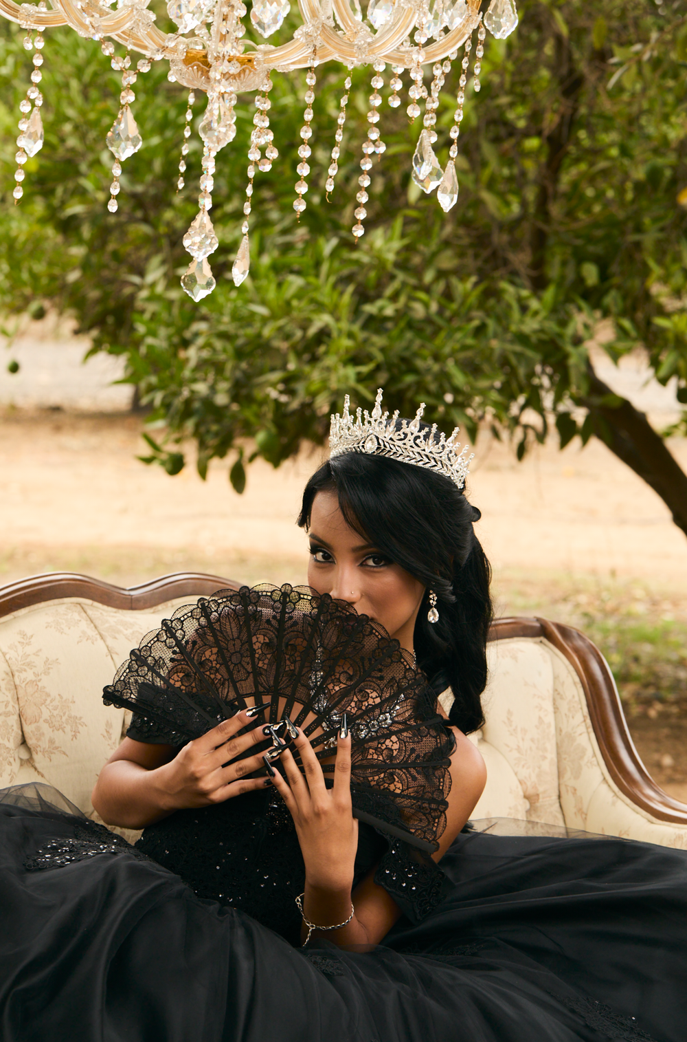 A woman in a black gown and crystal tiara holds a lace fan while seated on a vintage sofa outdoors.