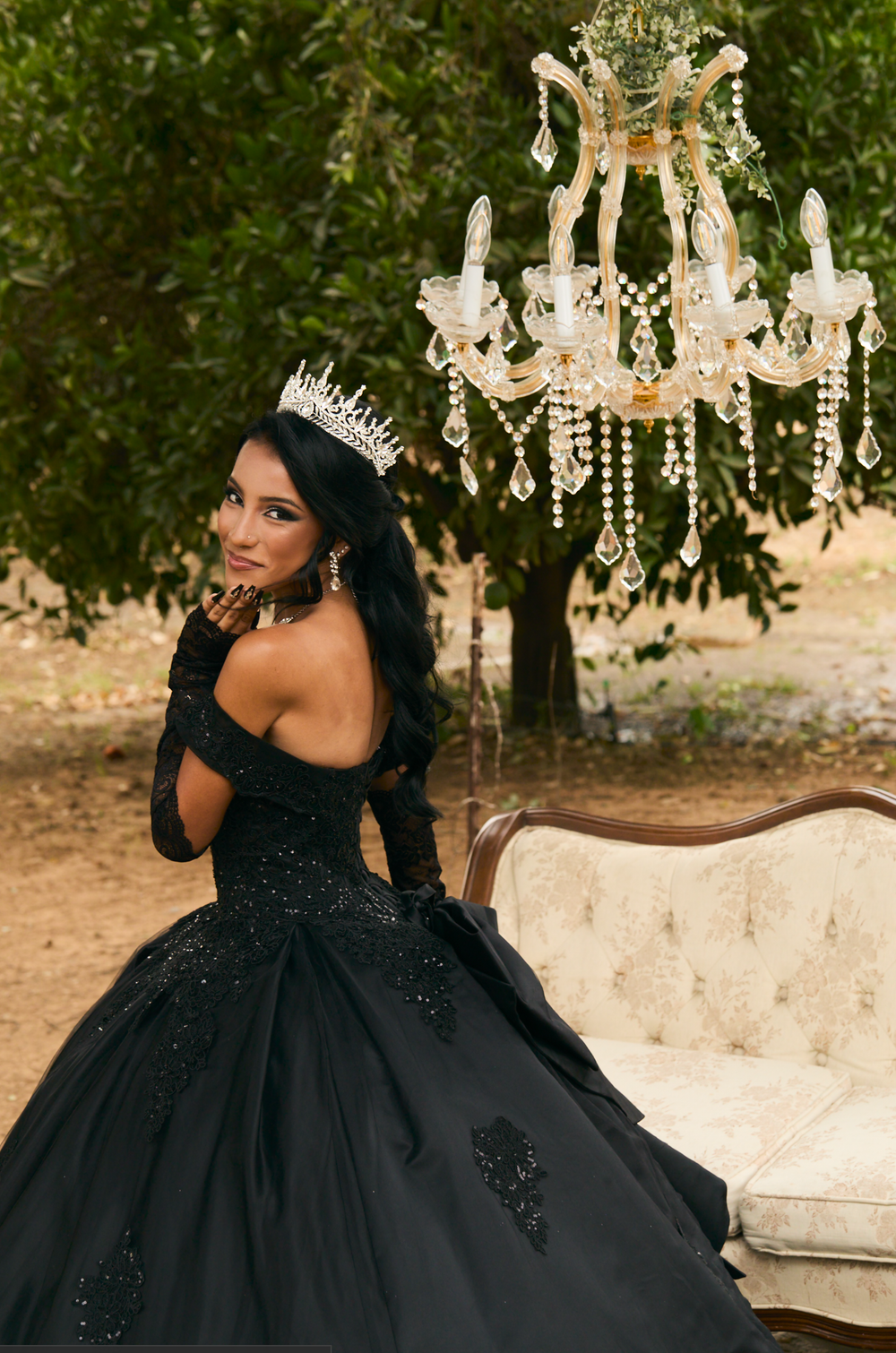 Woman in black ball gown and crown poses elegantly beside vintage sofa under crystal chandelier outdoors.