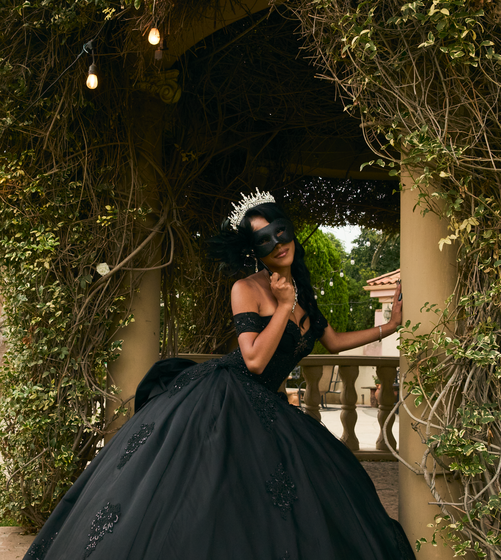 Woman in black ball gown with masquerade mask and tiara poses under vine-covered archway.