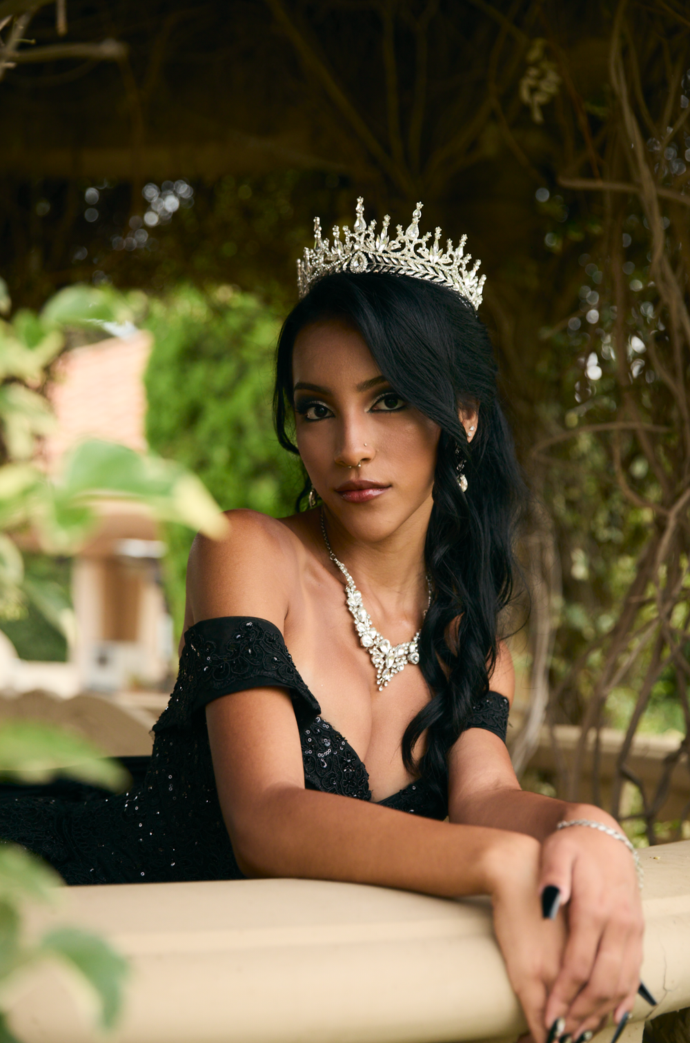 Young woman in black gown and crystal crown poses elegantly outdoors in a lush garden setting.