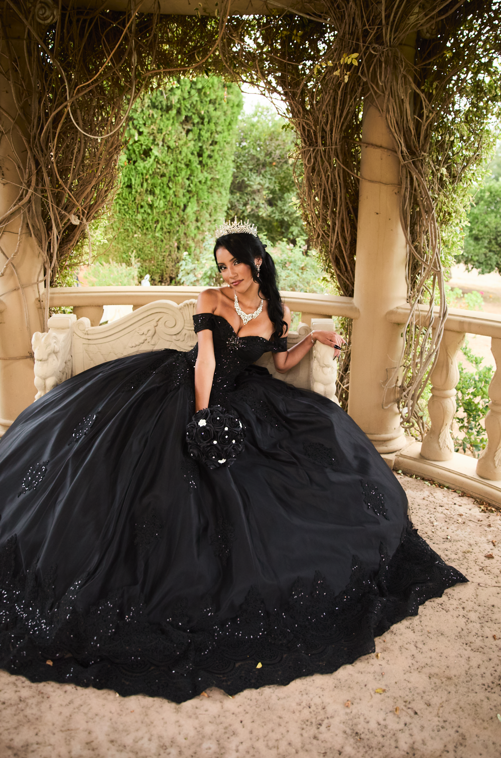 Woman in elegant black ball gown with tiara poses on ornate balcony surrounded by lush greenery.
