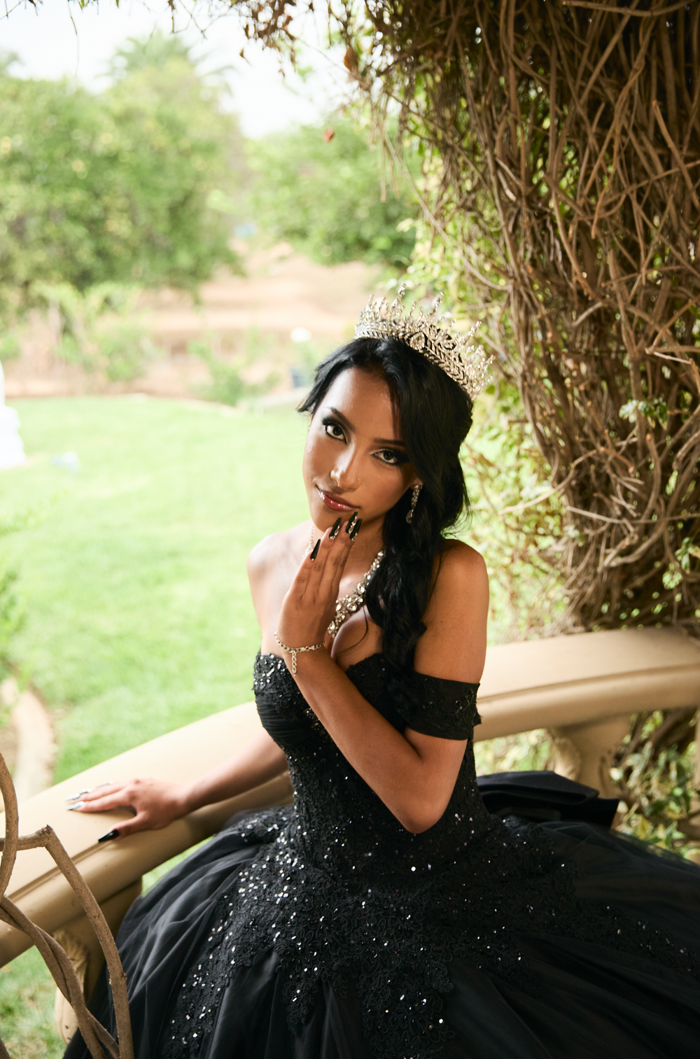Young woman in black sequin ball gown and gold tiara poses gracefully outdoors near lush greenery.