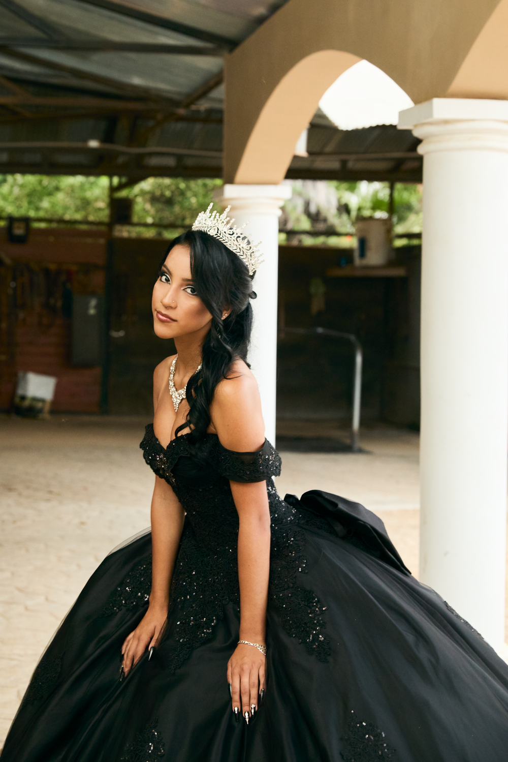 Young woman in black ballgown with gold crown poses elegantly by white column for quinceañera portrait.