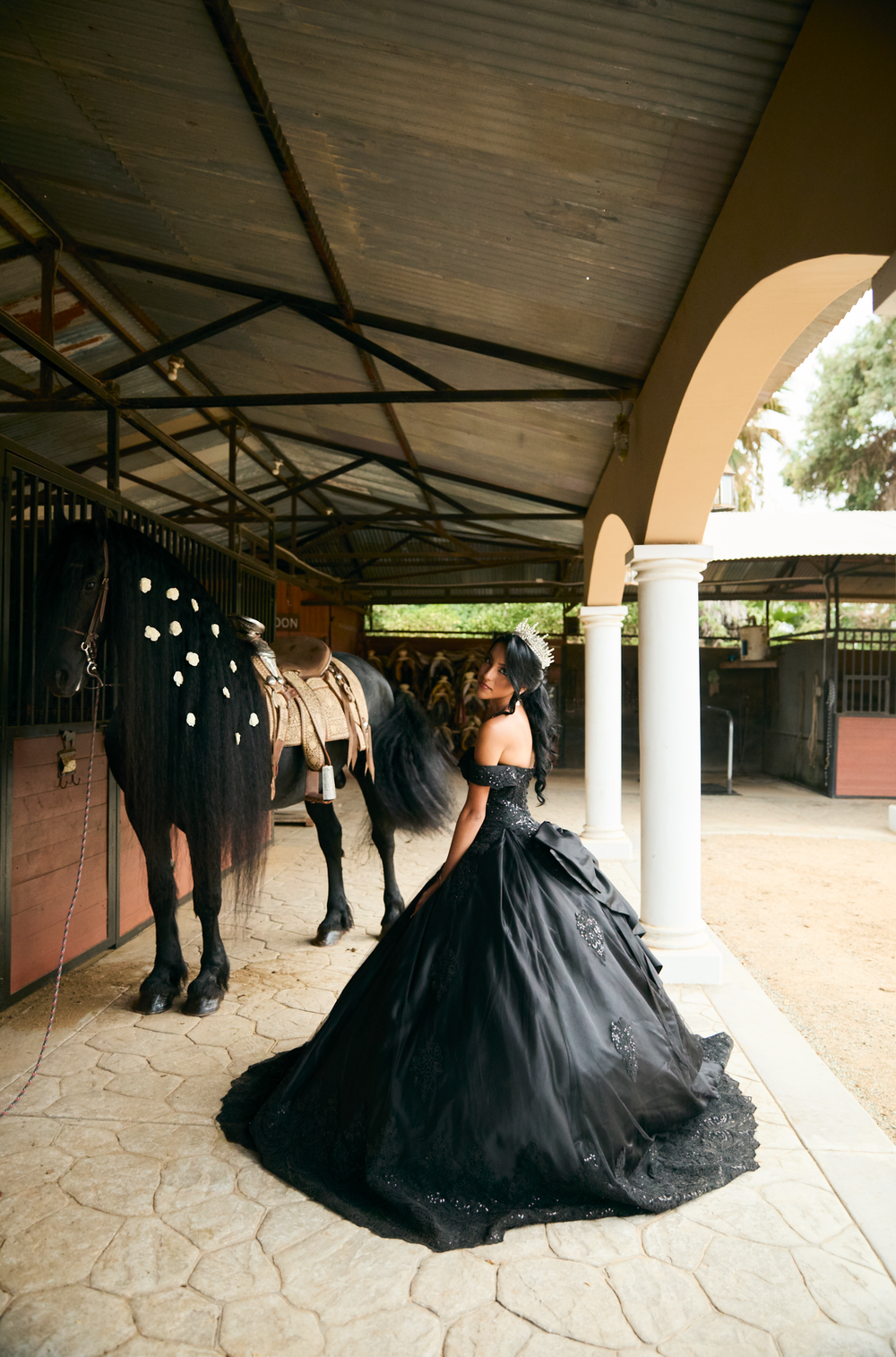 Woman in black ball gown stands beside a decorated horse at an elegant stable with white columns.