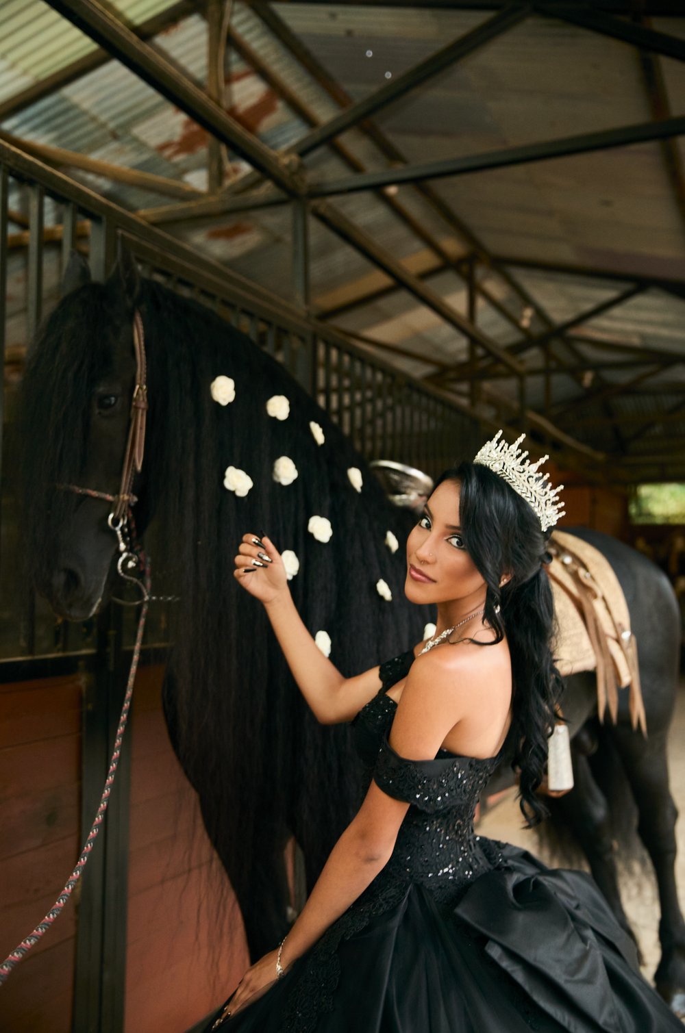 A woman in a black off-shoulder gown and tiara poses with a black horse adorned with white roses in a stable.