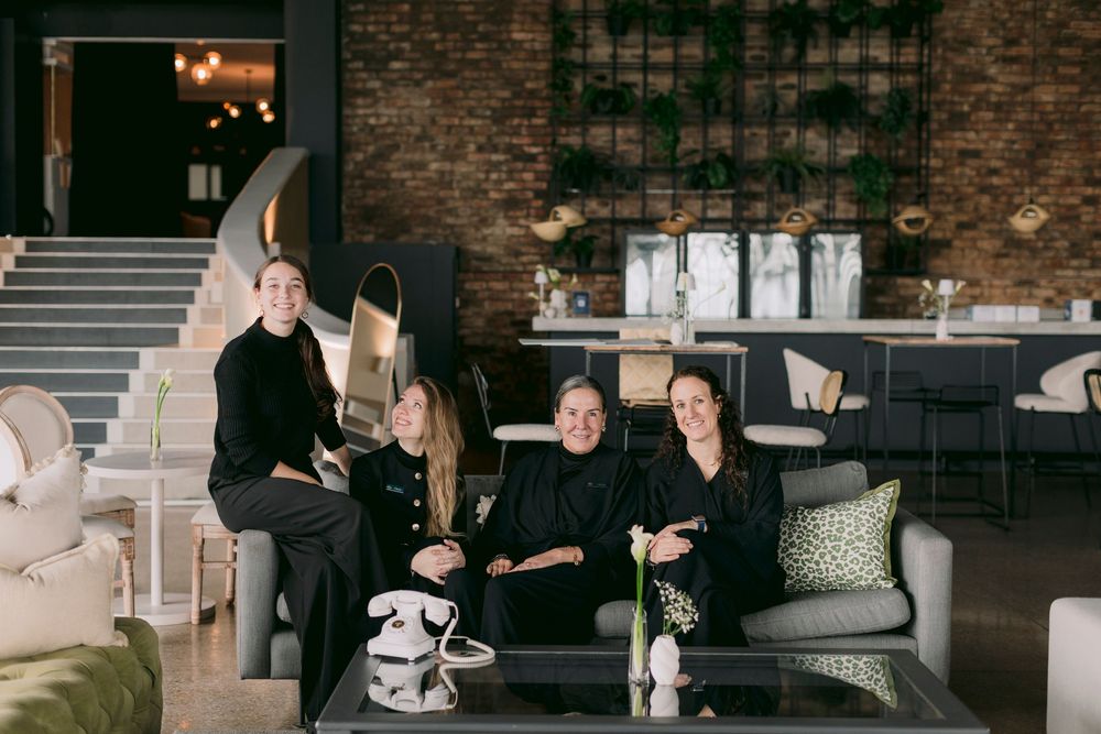 Four women in black outfits pose together in a modern, stylish lounge with green plant wall and brick accents.