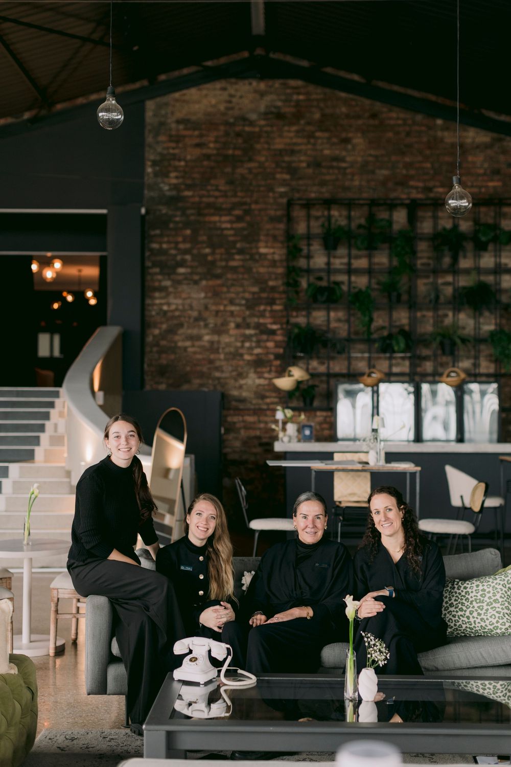 Four women in black outfits pose together in a stylish industrial restaurant with brick walls and green plants.