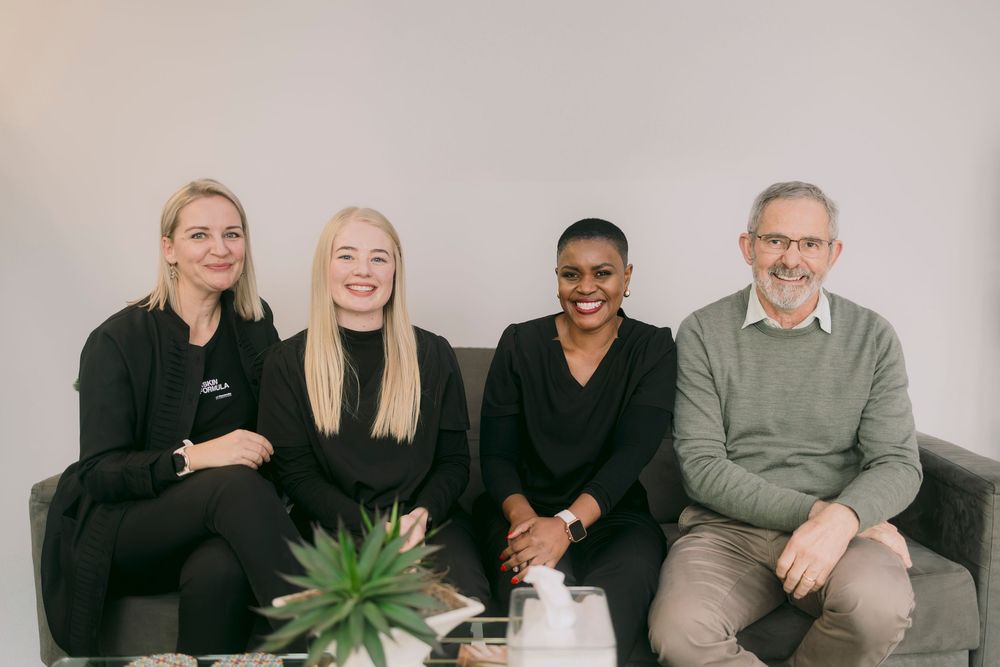 Four smiling professionals sitting together on a grey sofa in a modern office setting.