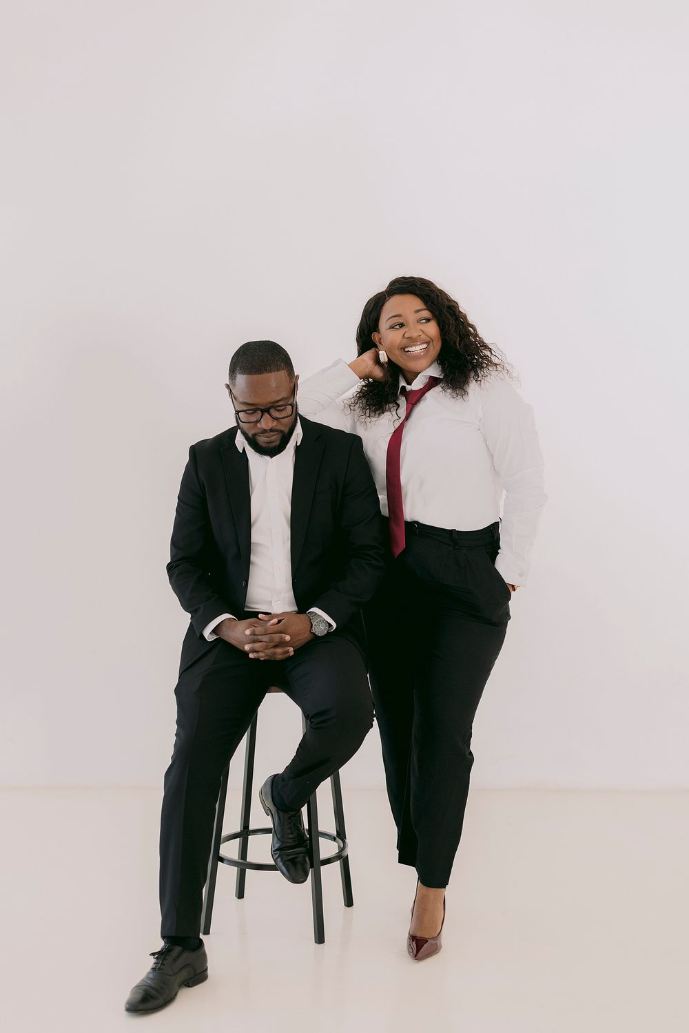 A man in a black suit sits on a stool while a smiling woman in a white shirt and red tie leans beside him.