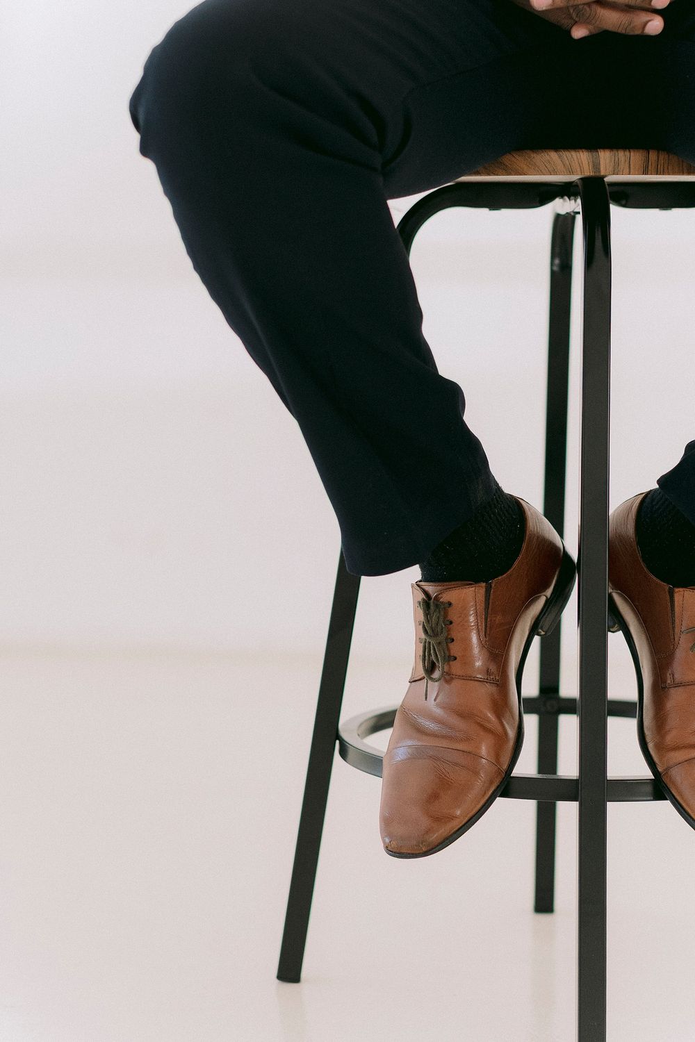 Person sitting on a black metal stool wearing dark trousers and brown leather lace-up dress shoes.
