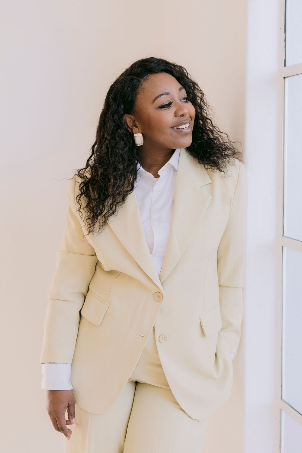 A smiling Black woman in a cream blazer suit and white shirt, with curly hair and gold earrings, posing by a wall.
