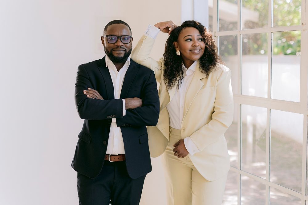 Two professionals in formal attire pose confidently near a bright window in a modern interior setting.