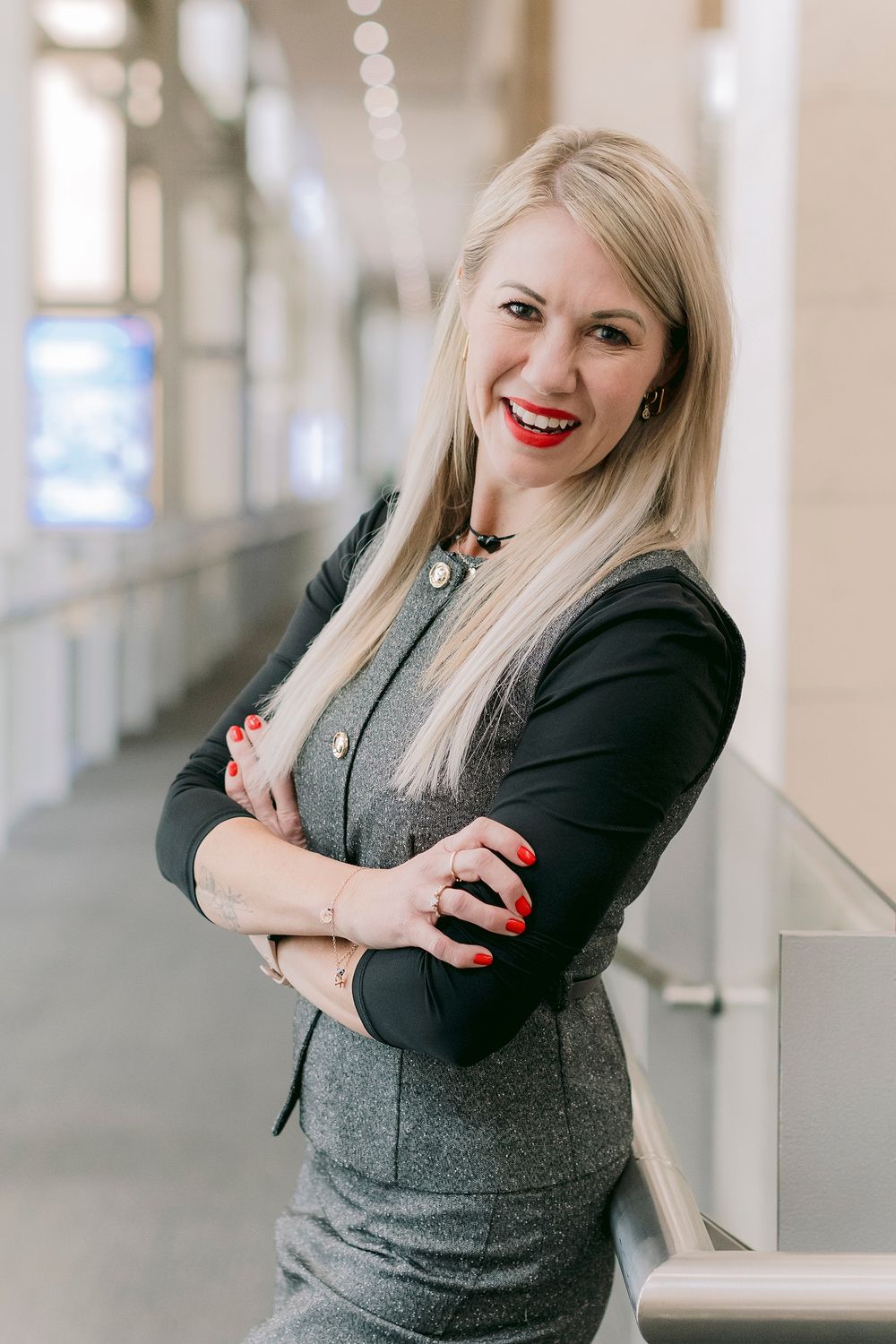 Professional blonde woman in gray blazer smiling confidently with arms crossed in modern office hallway.