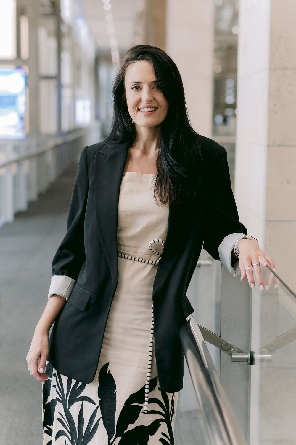 Professional woman in black blazer and floral skirt smiling in modern corridor, corporate portrait photography.