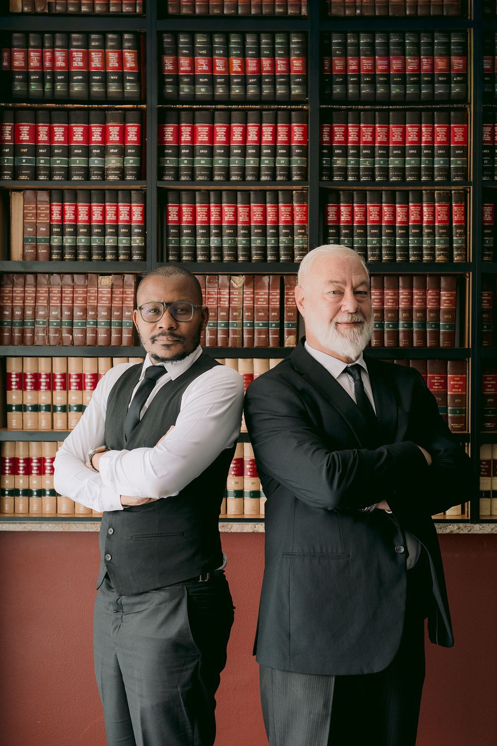 Two professional lawyers stand back-to-back with arms crossed in front of a law library bookshelf.
