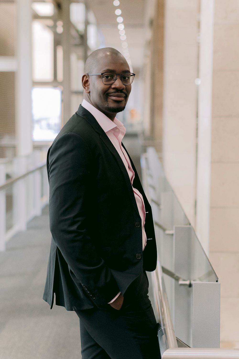 A professional Black man in a dark suit and pink shirt with glasses poses confidently in a modern building corridor.