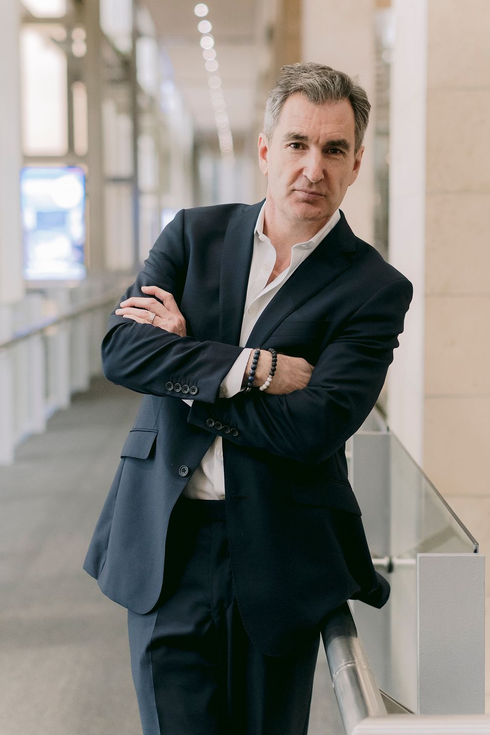Middle-aged businessman in navy suit stands with arms crossed in modern office corridor, exuding confidence.