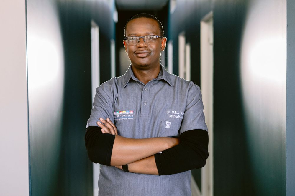 A smiling Black man with glasses stands confidently with arms crossed in a modern hallway wearing a gray button-up shirt.