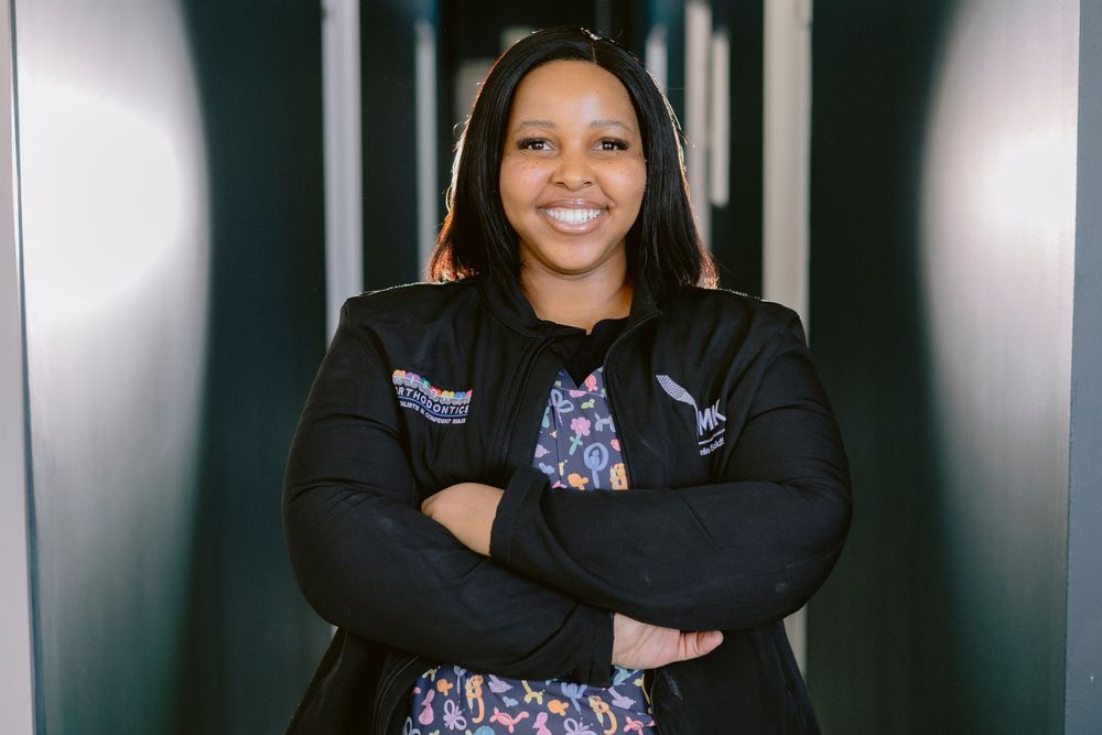Confident woman smiling with arms crossed, wearing a black jacket and floral top, standing in a modern corridor.