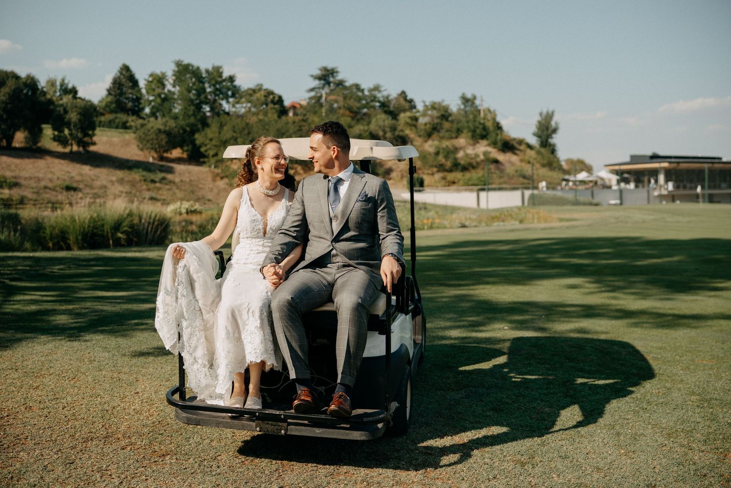 A bride and groom ride a golf cart together, smiling on a sunny golf course.