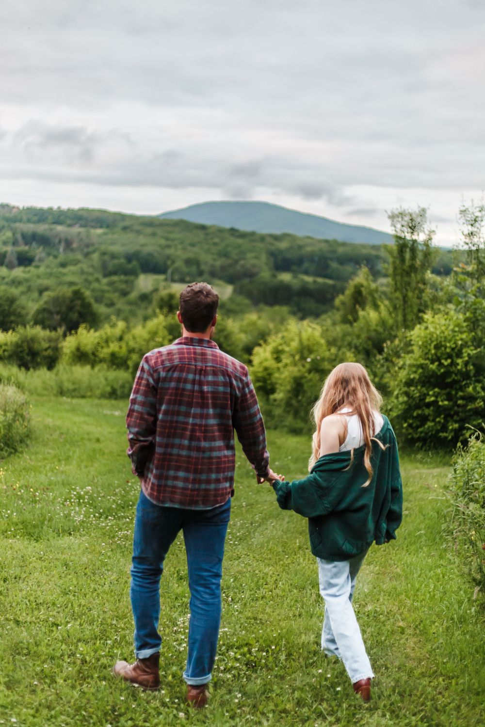 Couple holding hands walking through green meadow with mountain views and lush forest in background.
