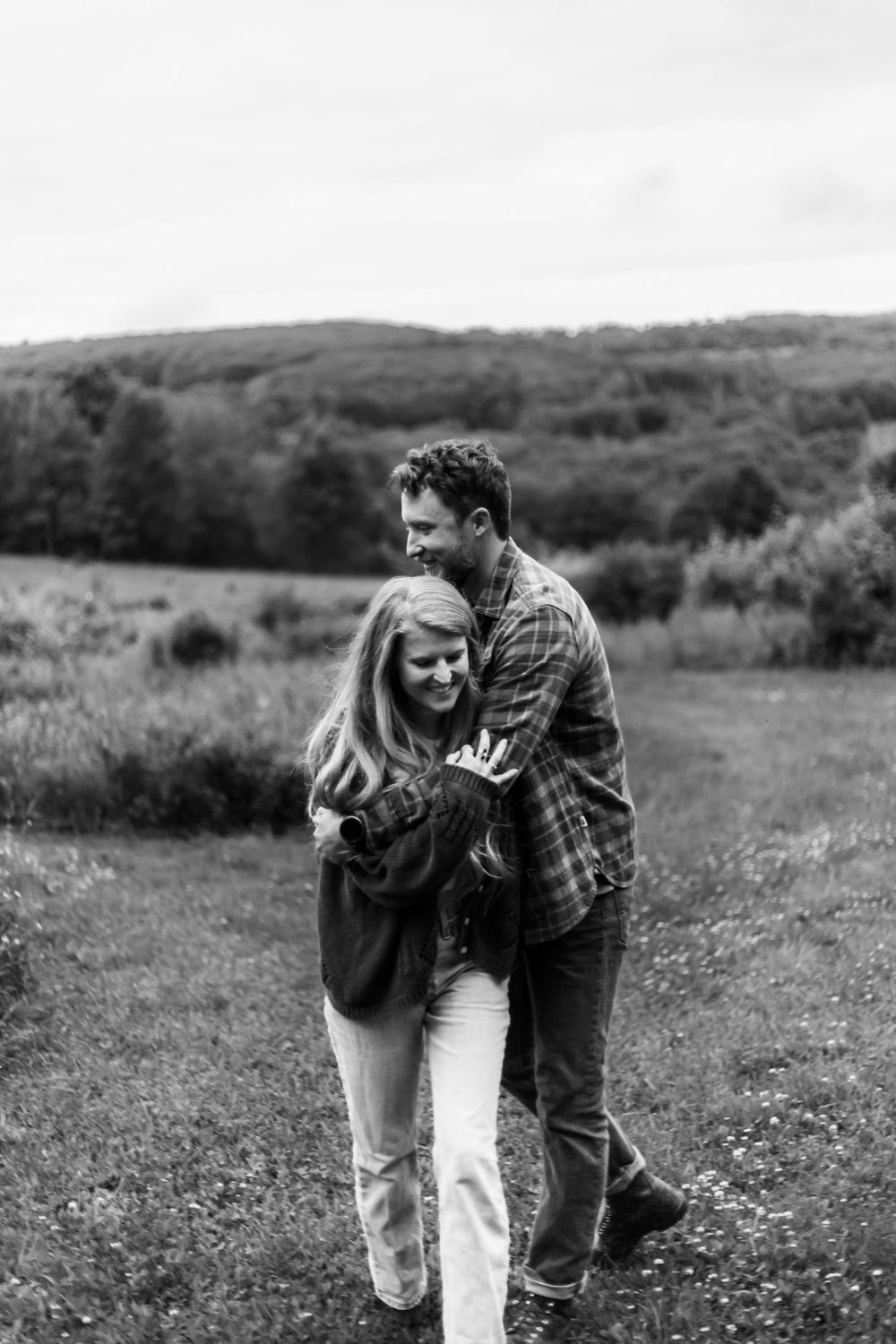 Black and white photo of a couple embracing and laughing on a grassy path with rolling hills in the background.