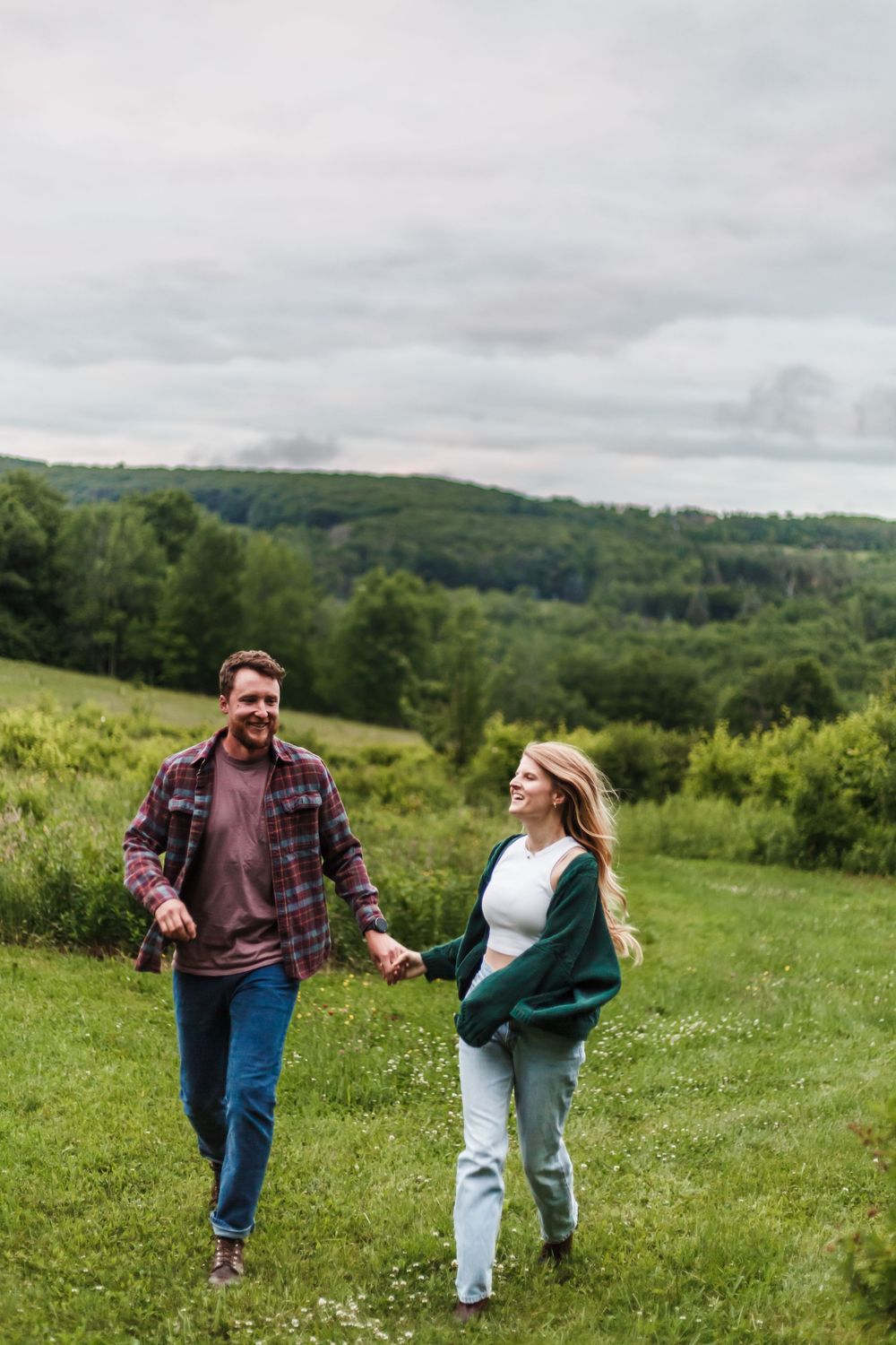 Couple holding hands walking through a lush green meadow with rolling forested hills in the background.