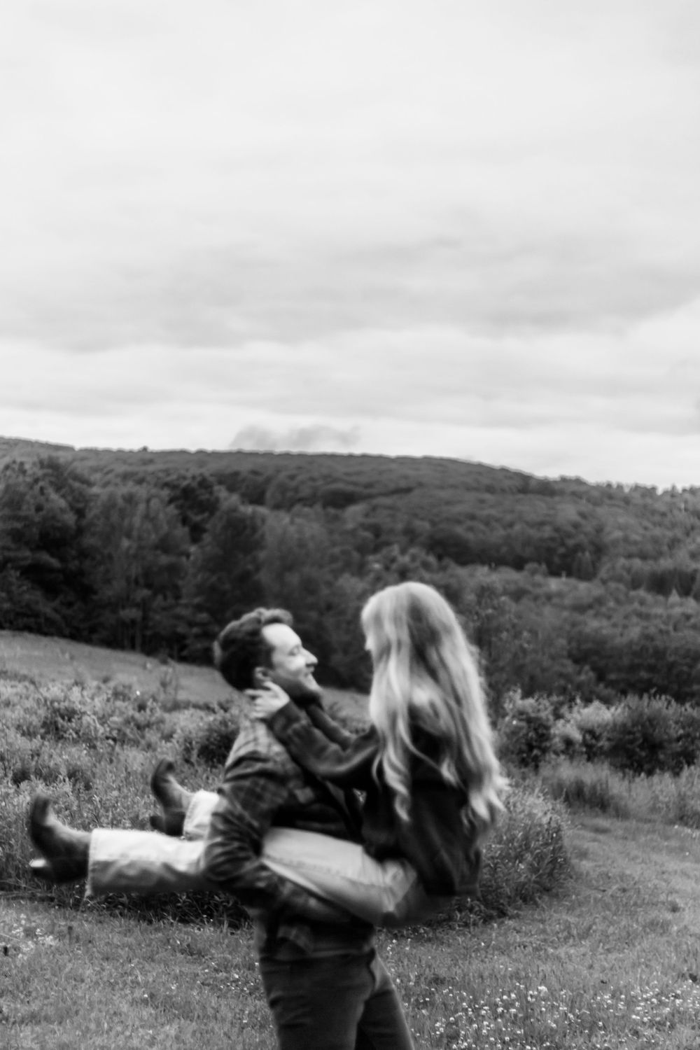 Black and white photo of a man lifting a woman in his arms outdoors with tree-covered hills behind them.