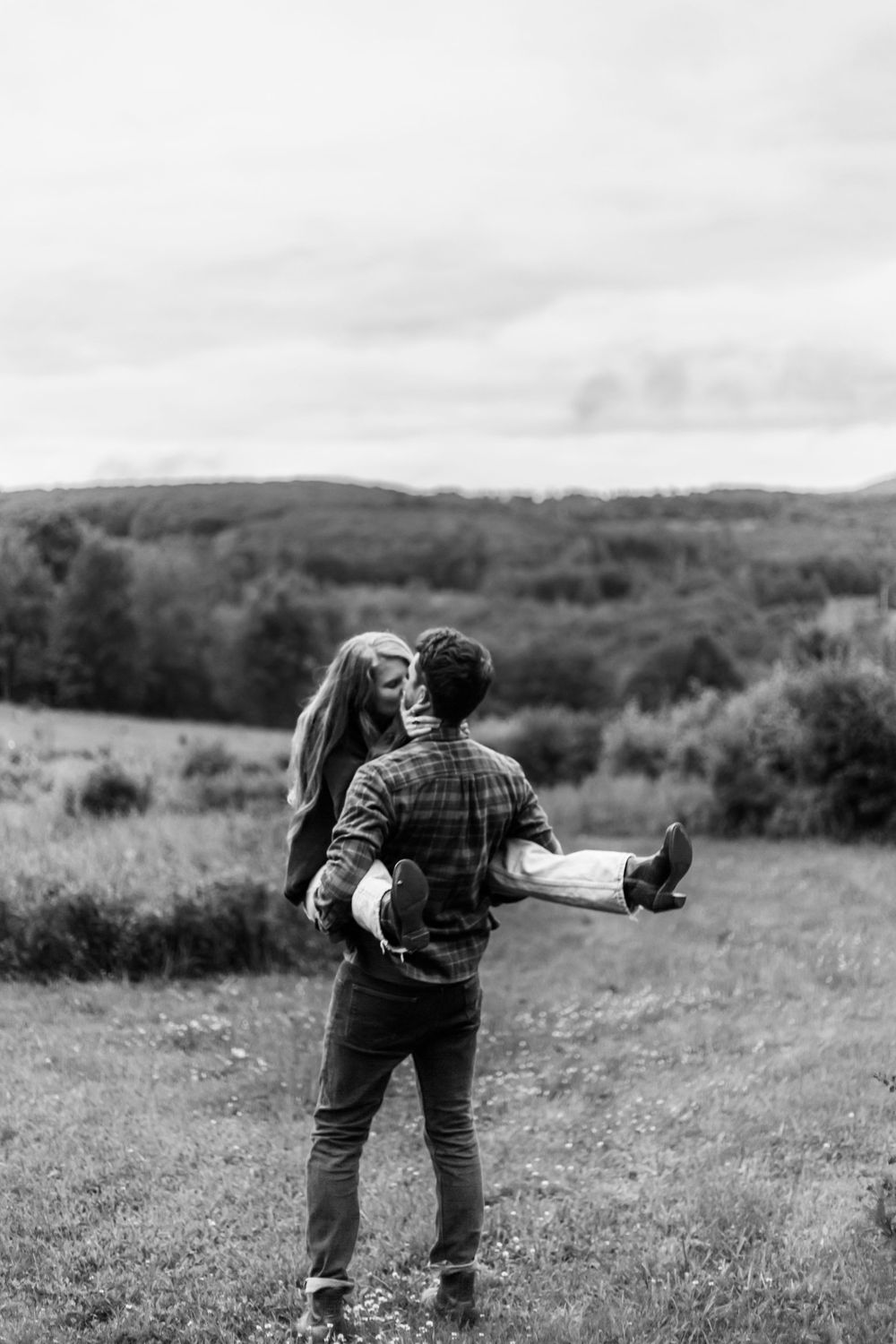 Black and white photo of a man carrying a woman in his arms in a scenic green field with rolling hills.