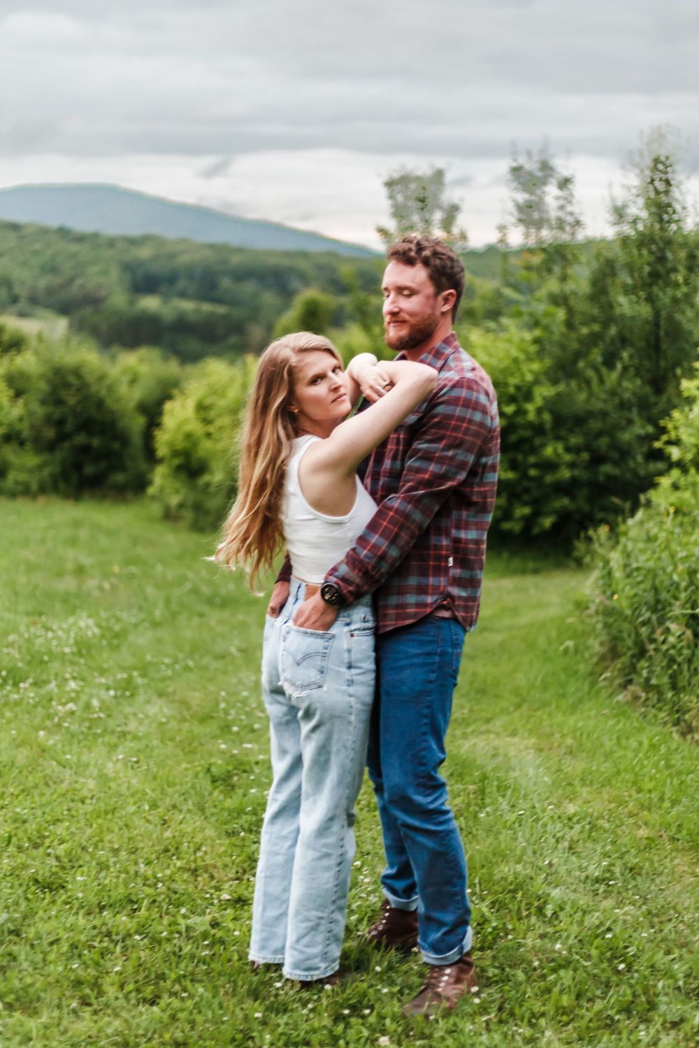 Couple embracing in a lush green field with mountain views, woman in white top and jeans, man in plaid shirt.