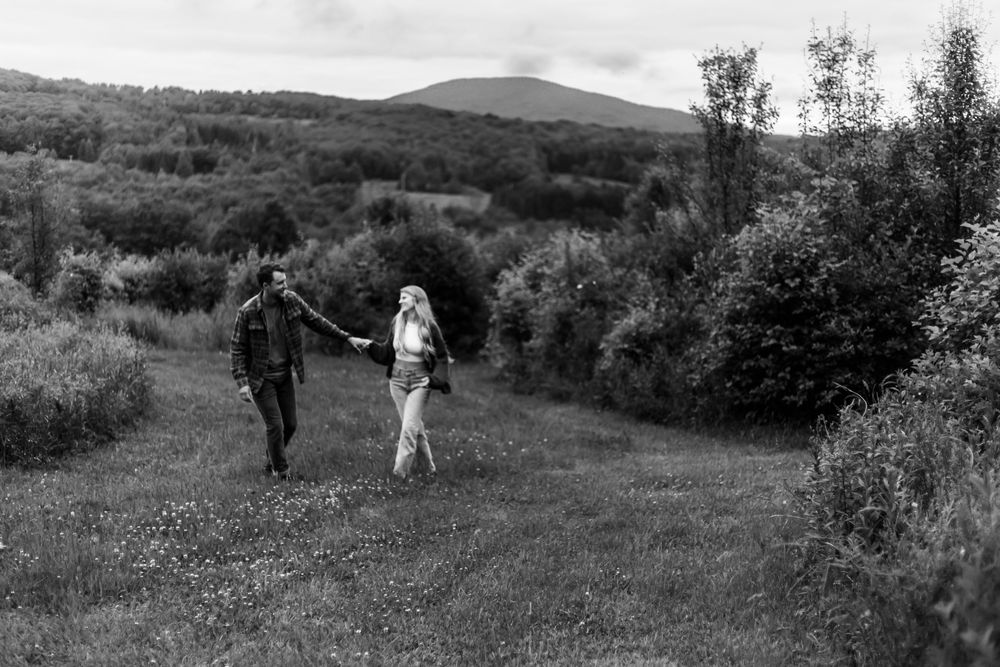 Black and white photo of a couple holding hands walking through a lush green meadow with mountains in the background.