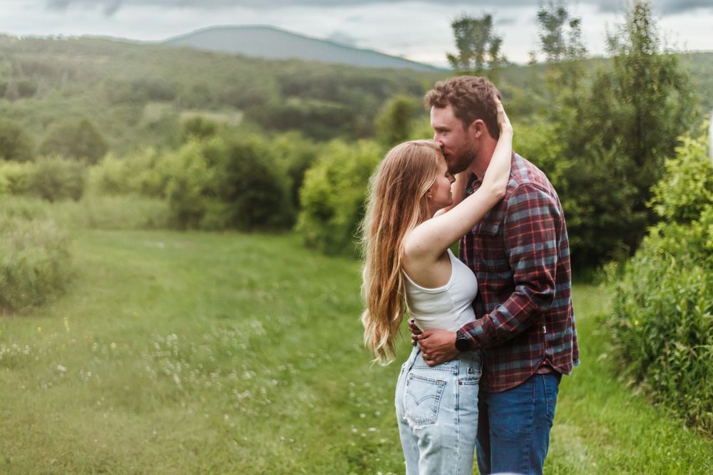 A couple embraces and nearly kisses in a lush green meadow with rolling hills and mountains in the background.